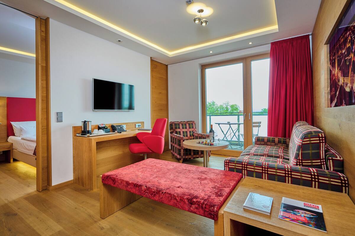 Quarto de hotel moderno com escrivaninha de madeira, uma cadeira vermelha, um sofá, televisão e varanda com cortinas vermelhas.