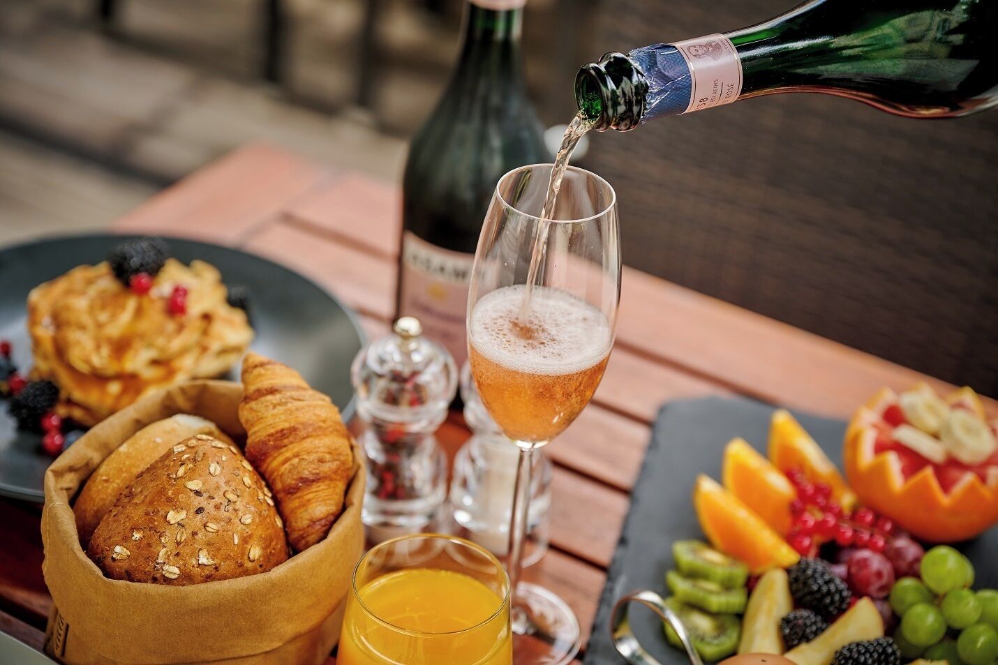 Une table dressée pour le petit-déjeuner avec des croissants, un verre de jus d’orange, une bouteille de champagne et une assiette de fruits.