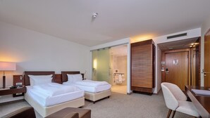 Habitación de hotel con dos camas, un escritorio, una silla y un baño visible a través de una puerta corrediza.