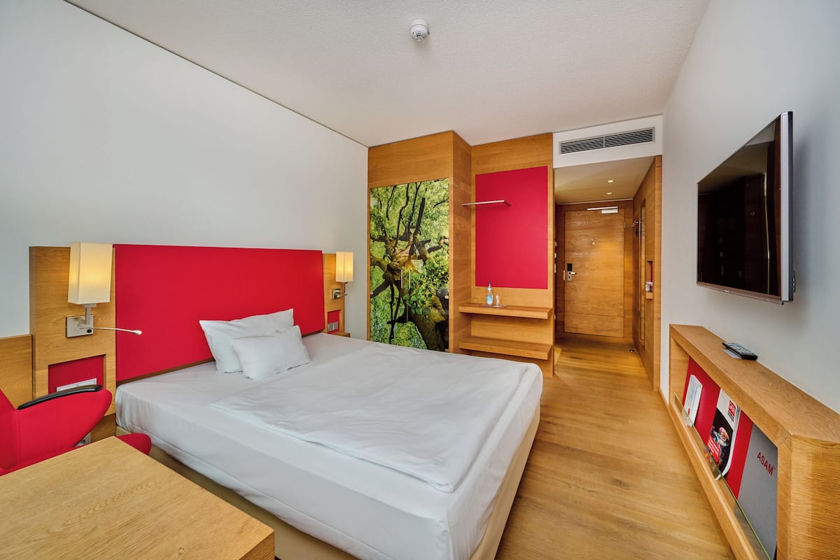 Quarto de hotel com uma cama grande, televisão, escrivaninha e uma parede com detalhes em vermelho.