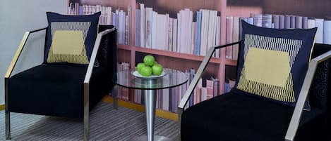 Two black chairs with yellow and black patterned cushions, a glass table with a metal base, and a bookshelf filled with books behind them.