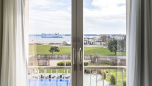 A view from a balcony with a pool, a ship in the distance, and a grassy area with a fence. - A-ROSA Travemünde (Lübeck)