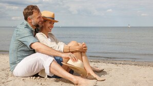 A couple sitting on a sandy beach with a calm sea and a ship in the distance. - A-ROSA Travemünde (Lübeck)