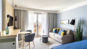 A modern hotel room with a sofa, a small round table, a desk with a chair, a bed, and a large window with curtains.