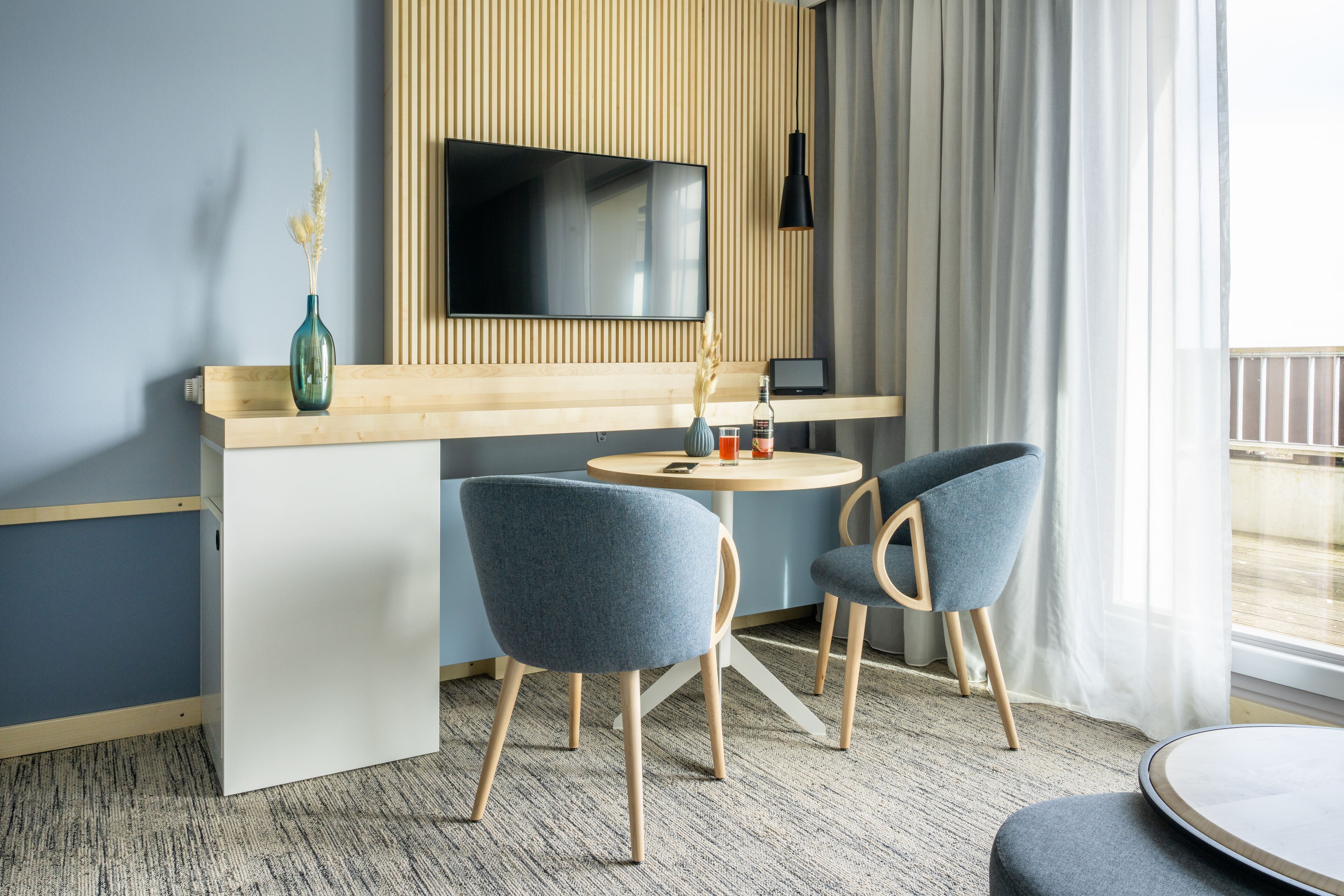 Una habitación de hotel moderna con un televisor de pantalla plana montado en la pared, un escritorio de madera, dos sillas azules y una mesa redonda.