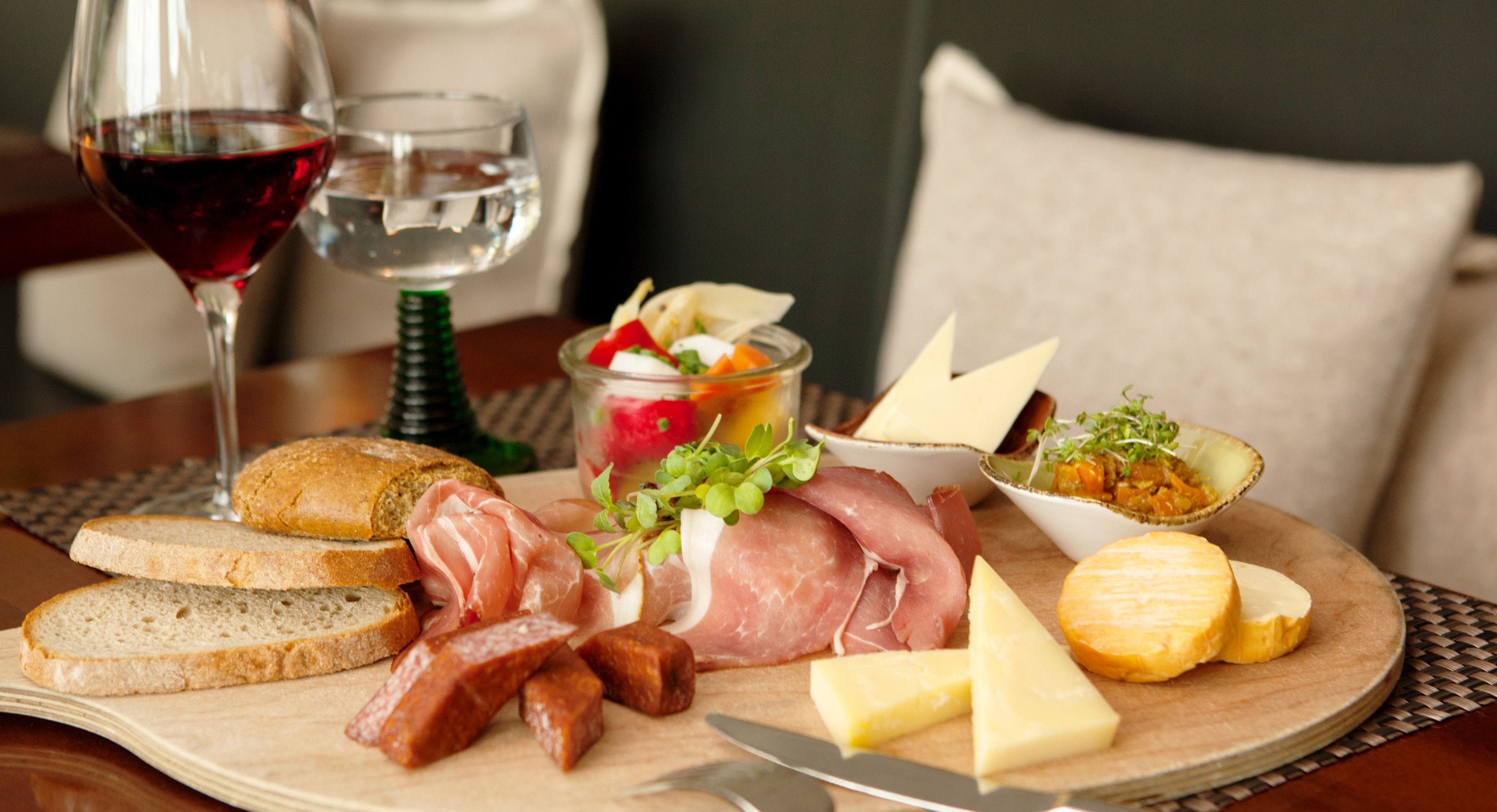 Ein Holzbett mit verschiedenen Käsesorten, Fleisch und Brot, begleitet von einem Glas Rotwein und einem Glas Wasser.