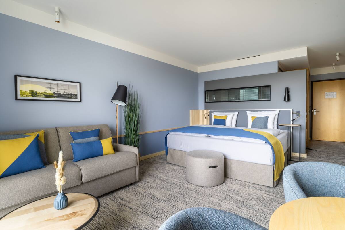 Una habitación de hotel moderna con cama, sofá y sillón, con una paleta de colores neutros y un ventanal grande.