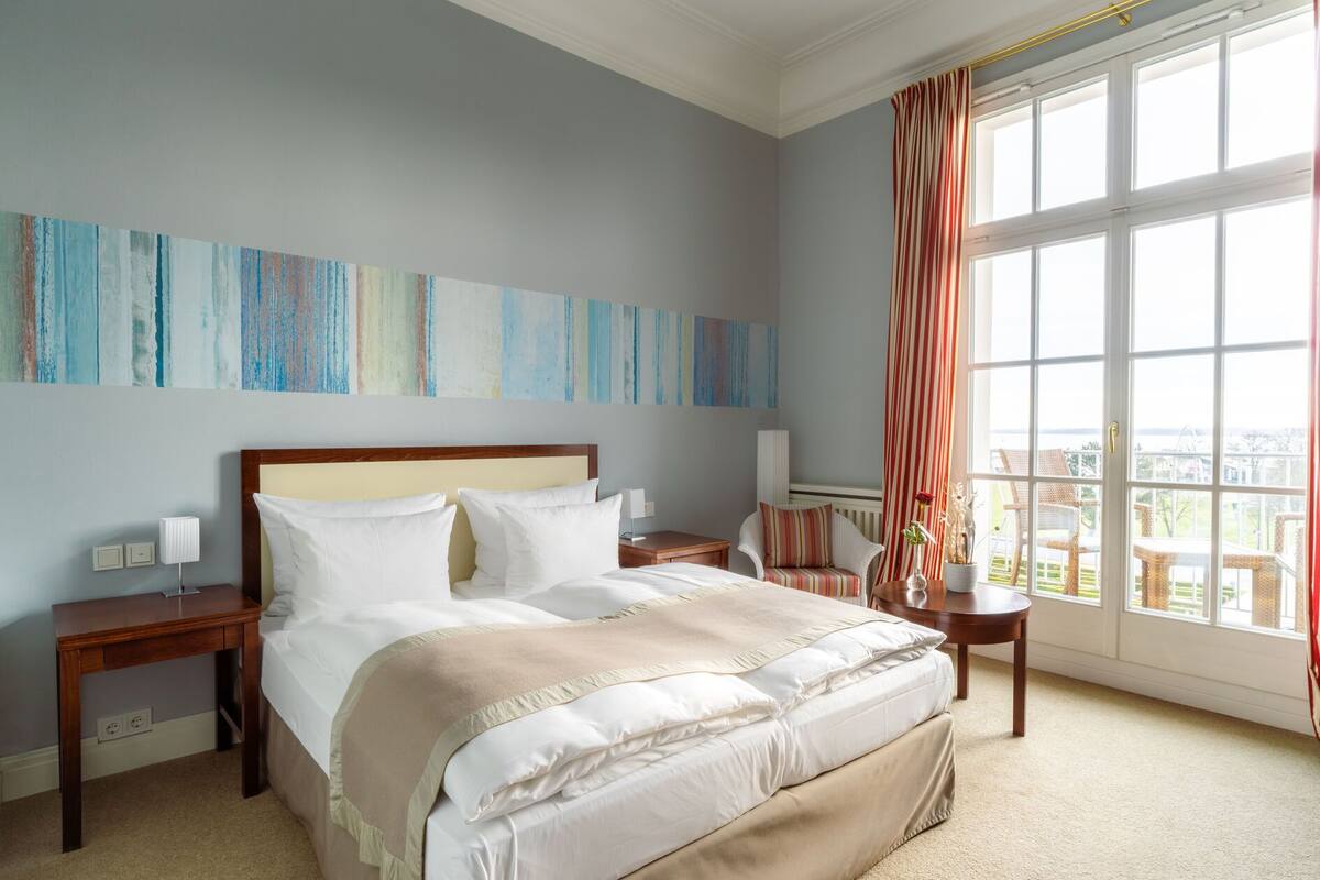 Habitación de hotel con una cama grande, una mesita de noche, una silla y un ventanal con cortinas.