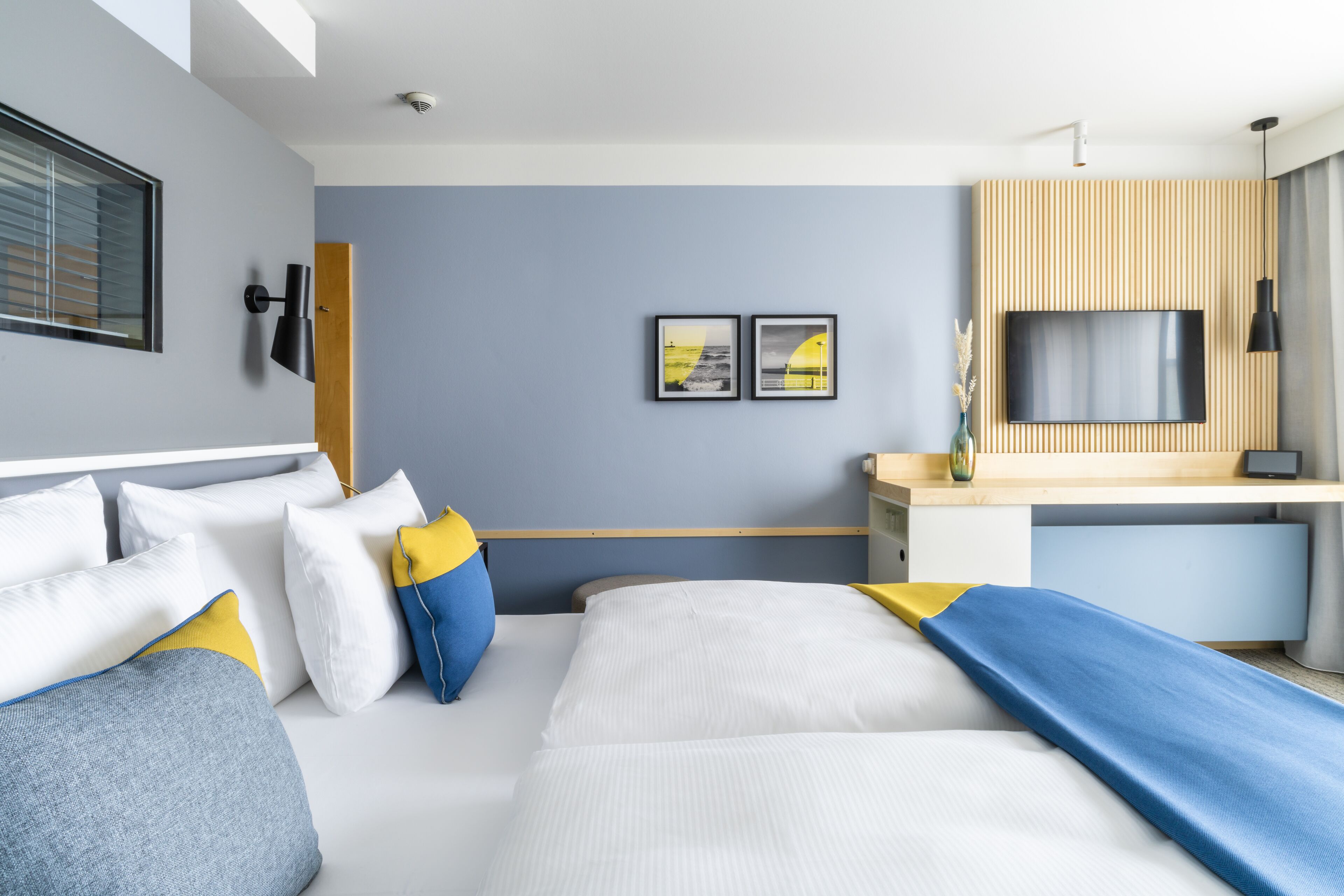 A modern hotel room with a bed, a desk, a TV, and artwork on the wall.