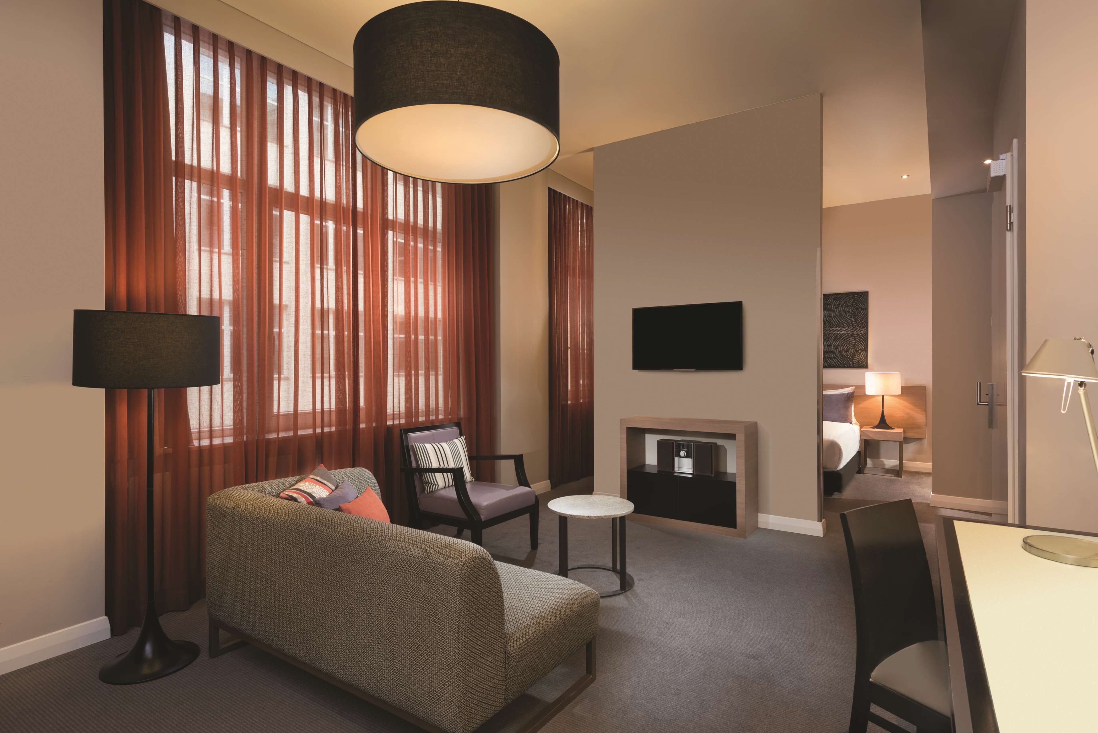 A hotel room with a sofa, armchair, small round table, and a flat-screen TV mounted on the wall.