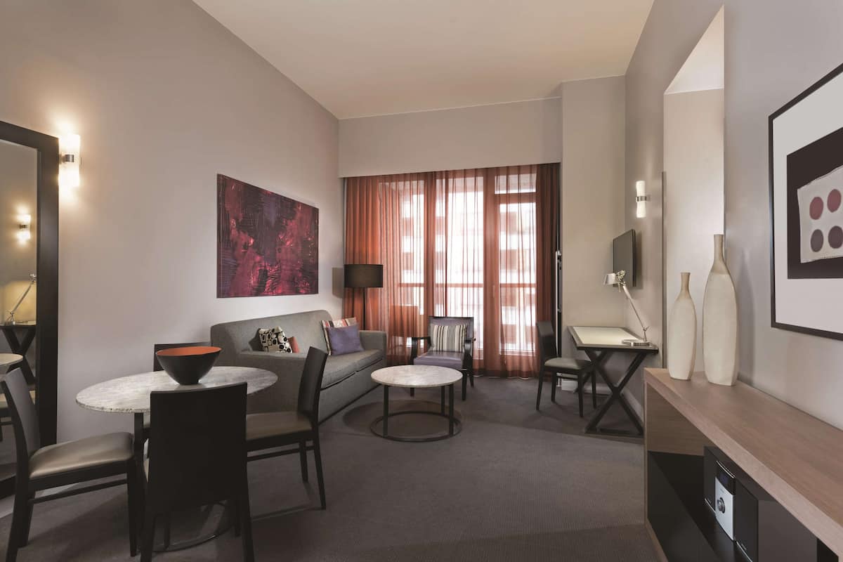 A modern hotel room with a sofa, two armchairs, a small dining table, a TV, and a fireplace.