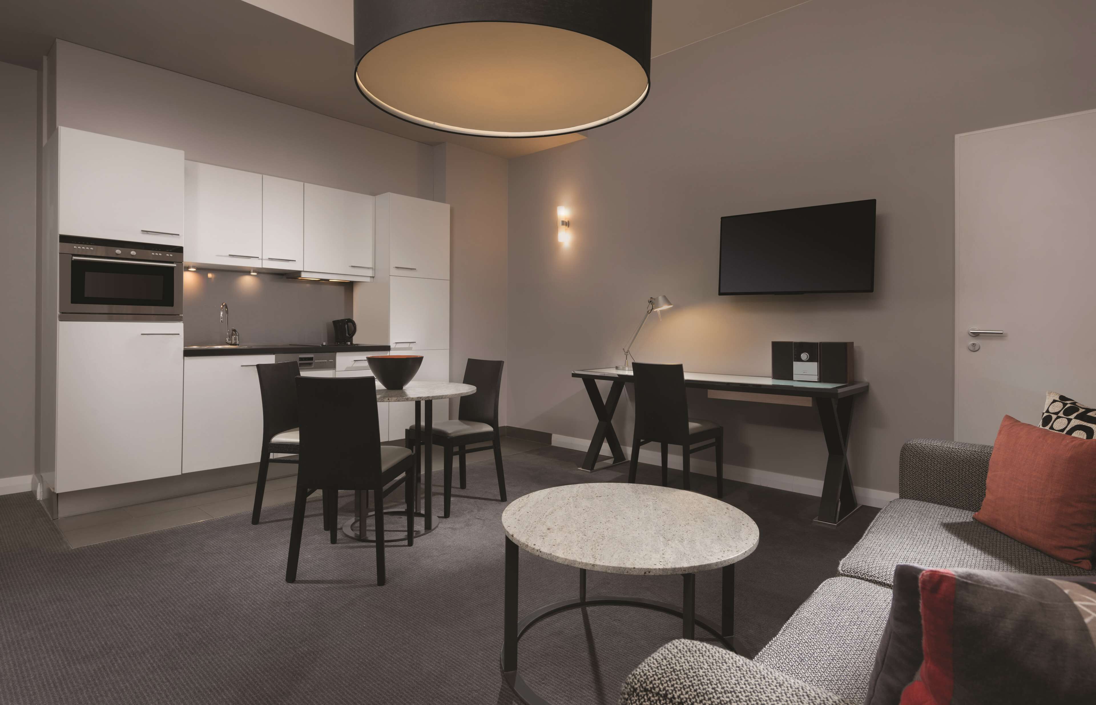 A modern kitchen with white cabinets, a dining area with a round table and chairs, a sofa, and a flat-screen TV mounted on the wall.