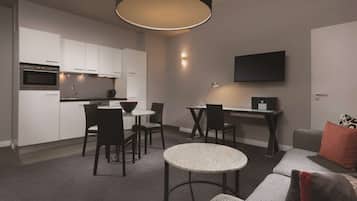 A modern kitchen with white cabinets, a dining area with a round table and chairs, a sofa, and a flat-screen TV mounted on the wall.