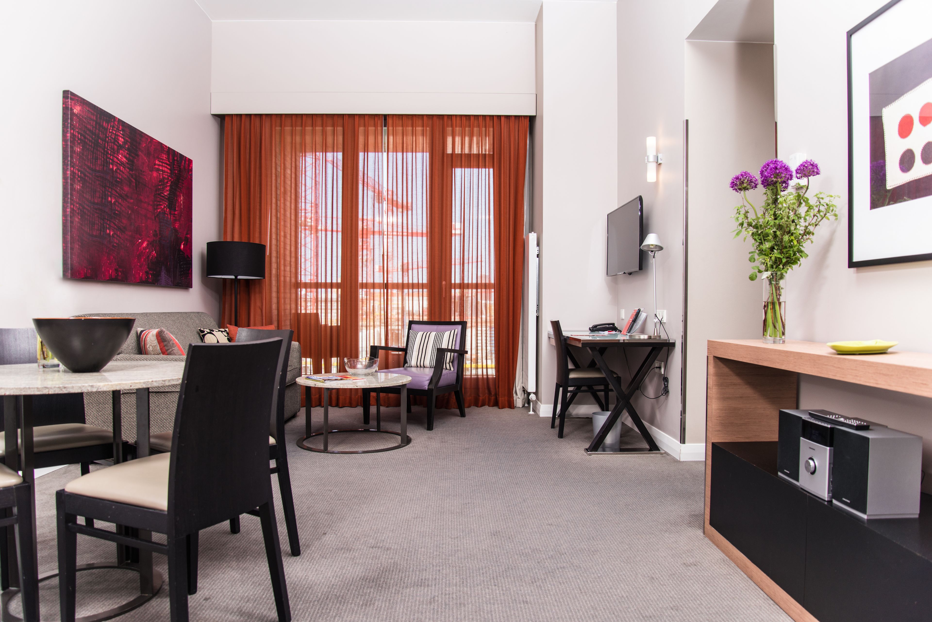 Ein modernes Hotelzimmer mit Essbereich, einem an der Wand befestigten Fernseher und einem großen Fenster mit Vorhängen.