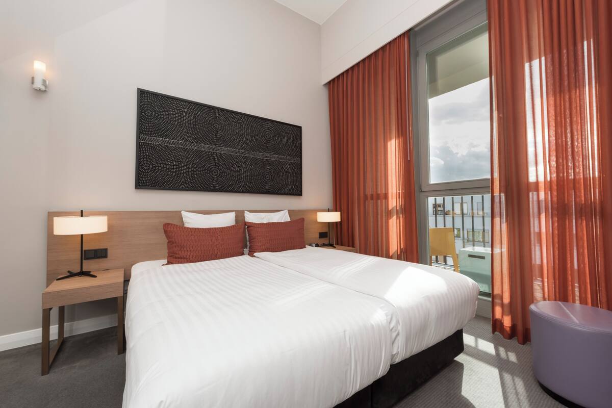 A hotel room with a large bed, two bedside tables with lamps, a large window with curtains, and a wall-mounted artwork.