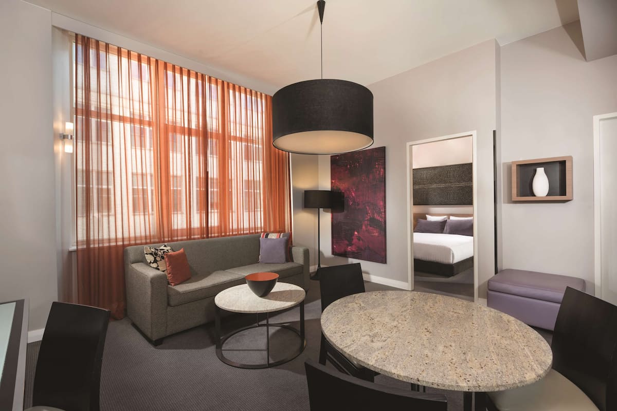A modern hotel room with a sofa, a round table, a bed, and a large window with curtains.