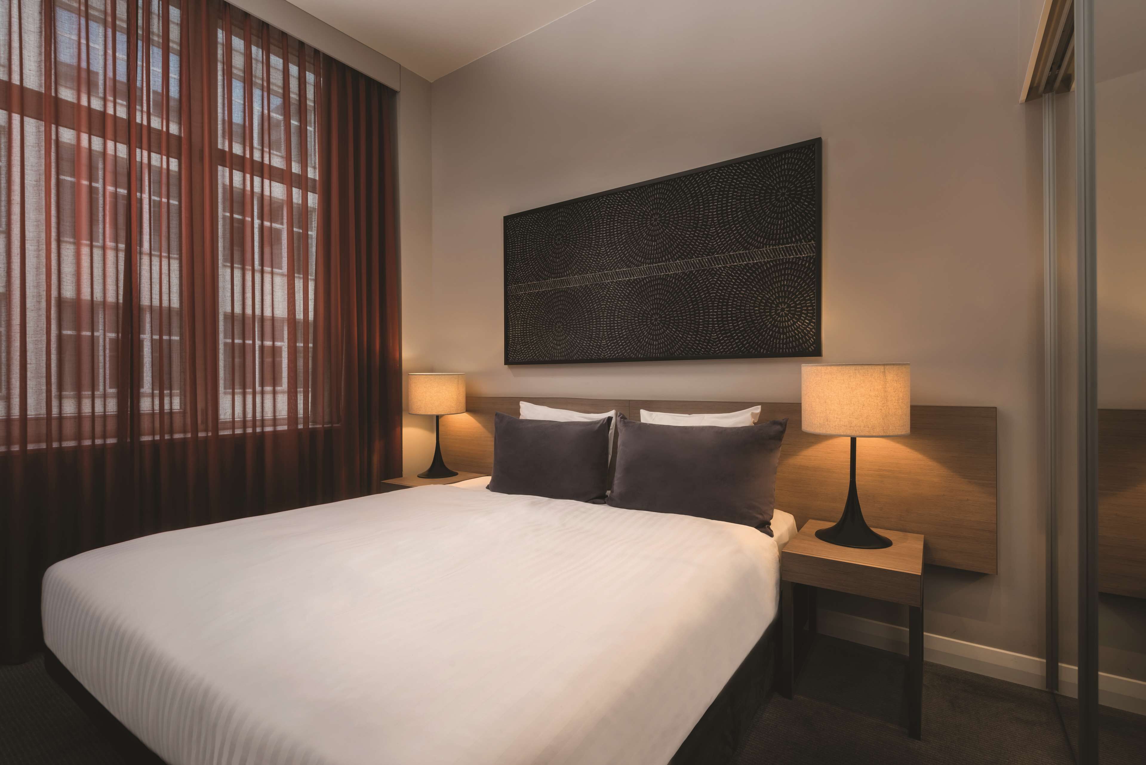 A hotel room with a large bed, two bedside tables with lamps, a wall-mounted artwork, and a window with blinds.
