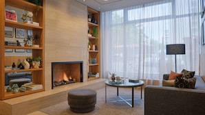 A modern living room with a fireplace, wooden shelving with decorative items, a round ottoman, and a large window with sheer curtains. - Adina Apartment Hotel Berlin CheckPoint Charlie (Berlin)