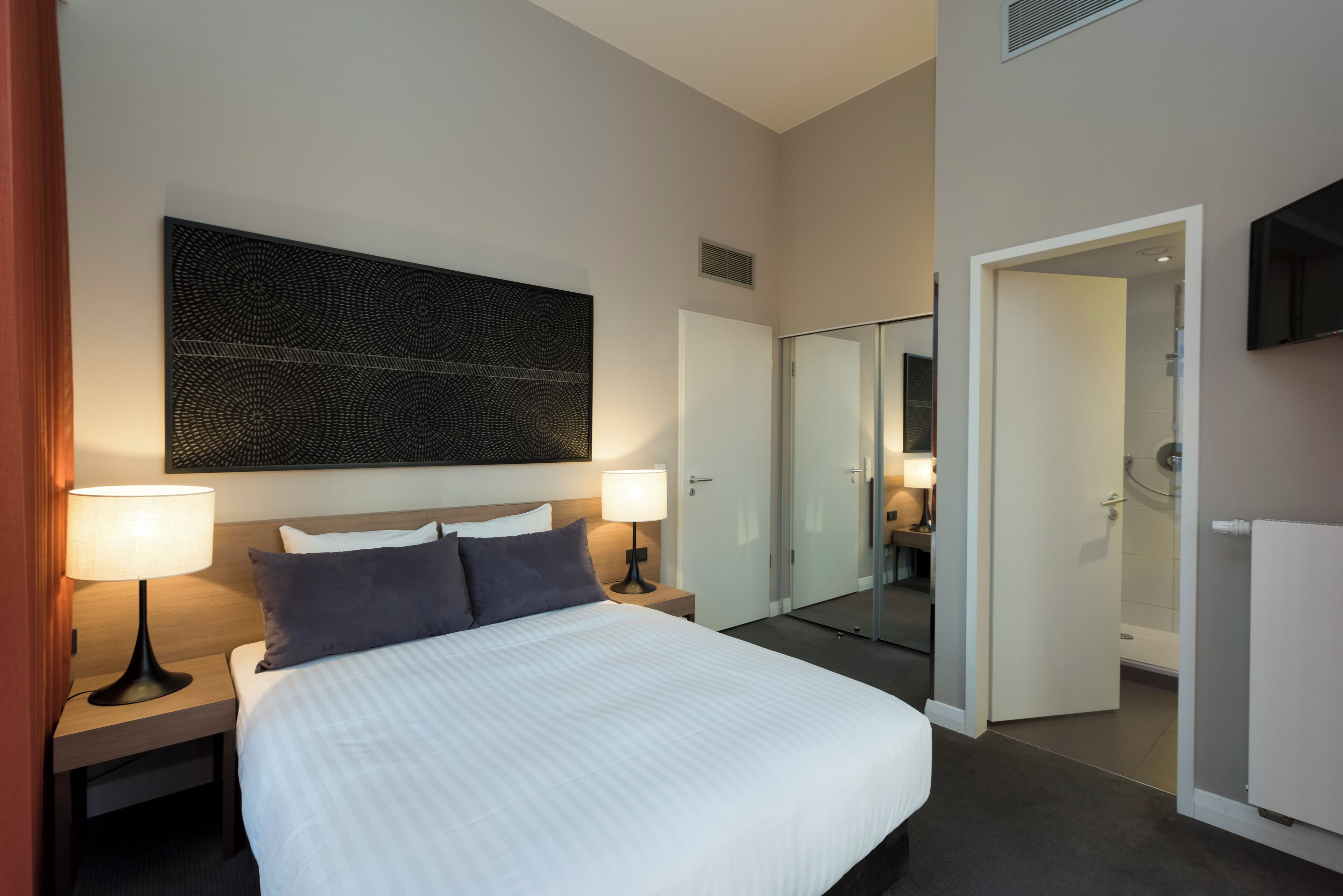 A hotel room with a large bed, two bedside tables with lamps, a wall-mounted artwork, a mirror, and a door leading to a bathroom.