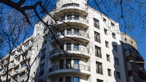 Un immeuble à plusieurs étages doté de balcons, une enseigne indiquant « Hôtel Volo », et plusieurs voitures garées devant.