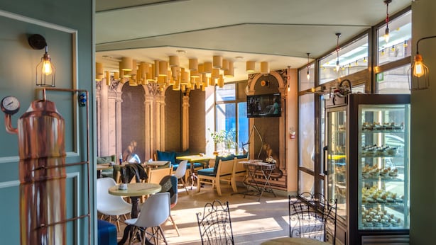 L’intérieur d’un restaurant se caractérise par de hauts plafonds, des luminaires suspendus, un espace central avec des tables et des chaises, et une vitrine où sont exposées des pâtisseries.