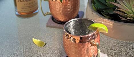 Un tavolo con una bottiglia di Tito's Handmade Vodka, due bicchieri di rame con cannucce, una fetta di lime e una piccola pianta grassa in vaso.