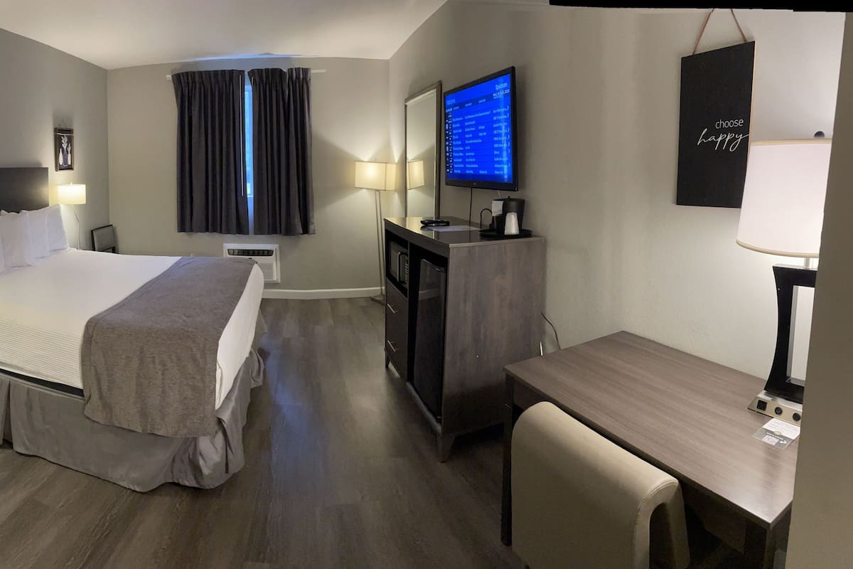 Habitación de hotel con cama, mesitas de noche, escritorio, silla, televisor y pantalla en la pared que muestra un menú.