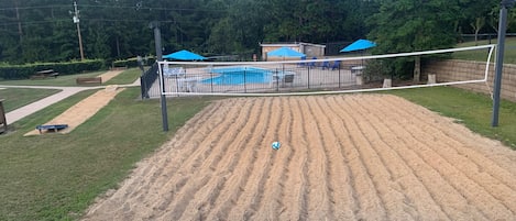 A sandy volleyball court with a fence, a pool, and blue umbrellas in the background.