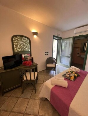 Une chambre d’hôtel avec un lit, un bureau avec une télévision, une chaise, une petite table avec un plateau de nourriture, et une salle de bain visible à travers une porte ouverte.