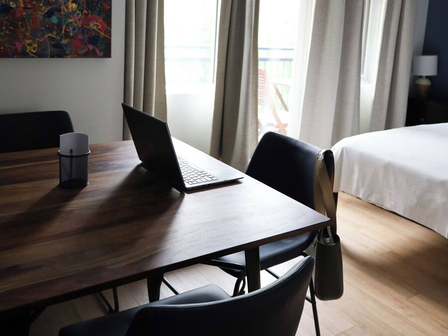 A hotel room with a wooden desk, a laptop, a black chair, a bed, and a window with curtains.