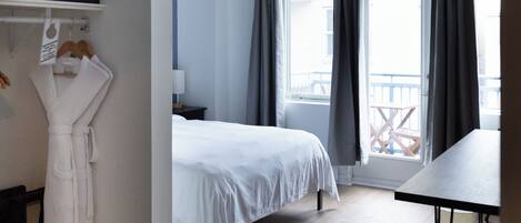 Ett hotellrum med en säng, ett skrivbord, en stol, en badrock som hänger på en klädställning och en balkong med gardiner.