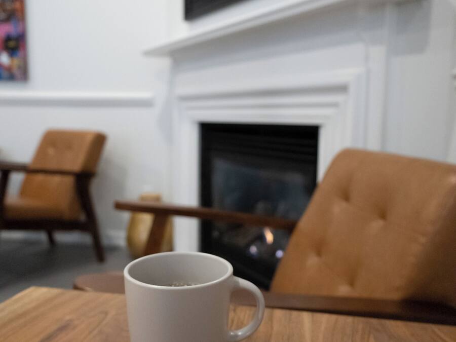 A white mug on a wooden table in a room with a fireplace, two brown leather chairs, and a framed picture on the wall.
