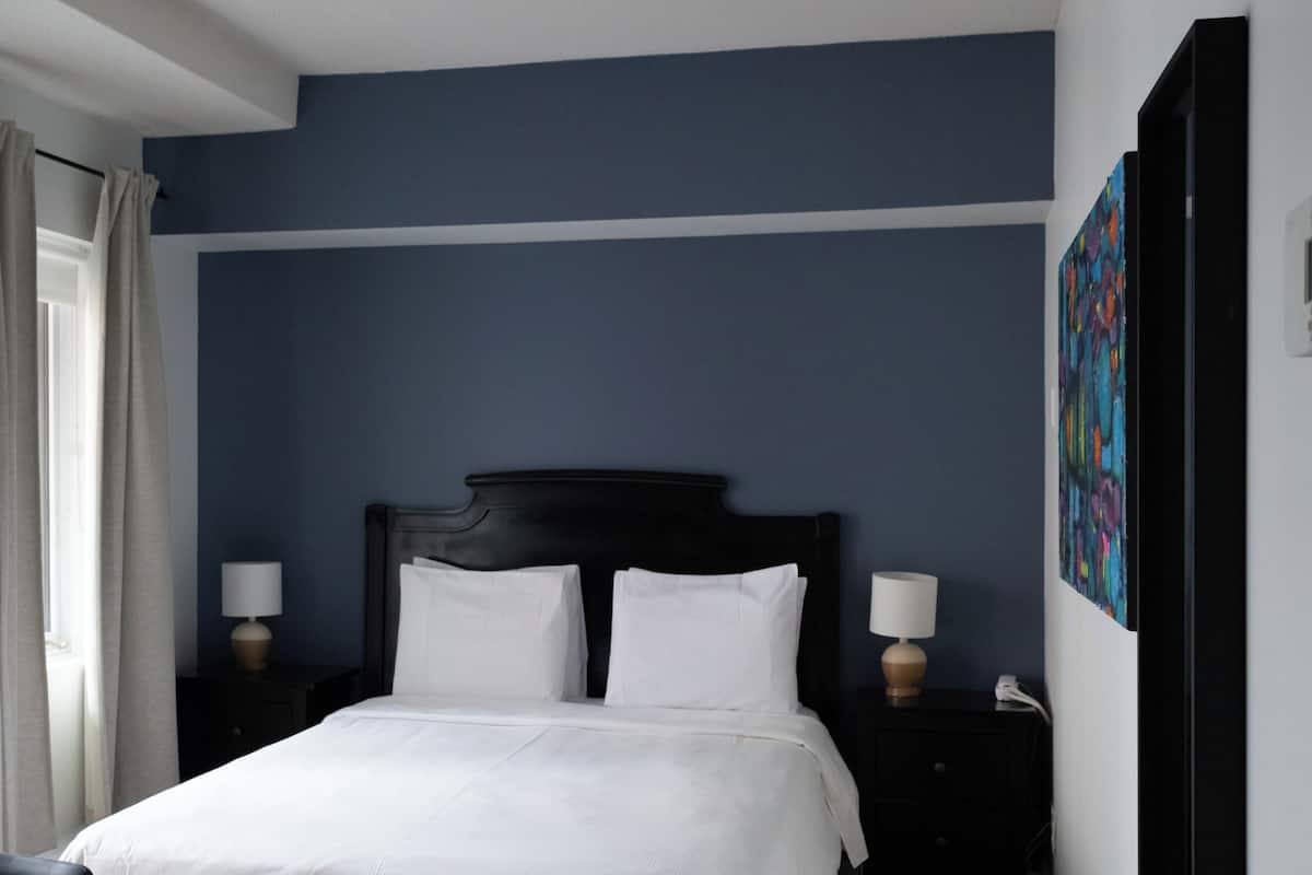 A hotel room with a large bed, a bedside table with a lamp, a chair, and a wall with a colorful painting.