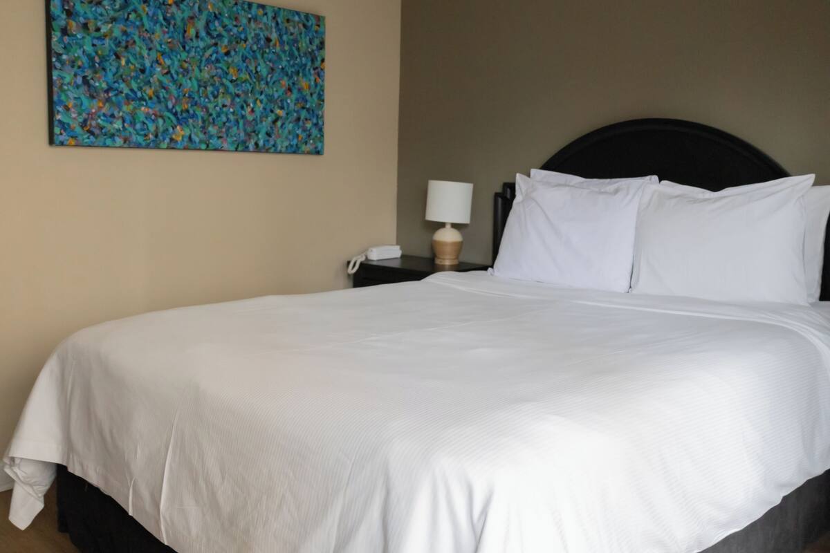A hotel room with a large bed, a nightstand with a lamp, and a colorful abstract painting on the wall.