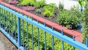 Un bâtiment en briques avec un balcon rouge et bleu, un parasol bleu et un jardin où poussent différentes plantes.