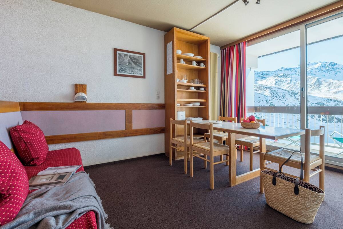 A cozy room with a wooden dining table, chairs, a bookshelf, a sofa with red cushions, and a view of a snowy mountain through a large window.