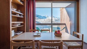 A dining area with a wooden table and chairs, a view of mountains through a large window, and a shelf with dishes. - Résidence Pierre & Vacances Val Thorens Le Gypaete (Les Belleville)