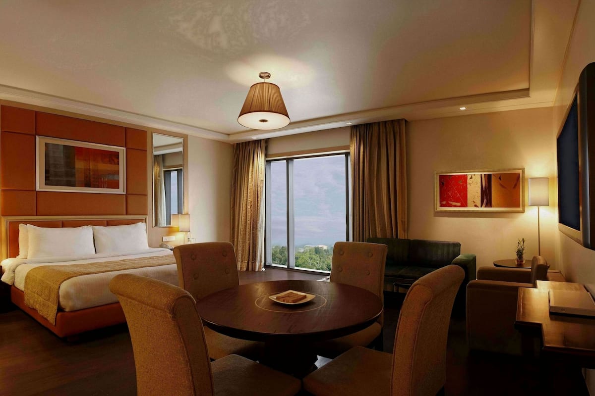 A hotel room with a bed, a dining table, chairs, a TV, and a large window.