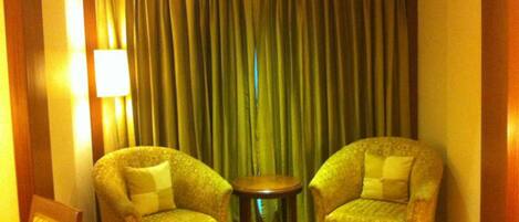 Una habitación con dos sillones, una pequeña mesa redonda, una lámpara y un cortinaje de fondo.