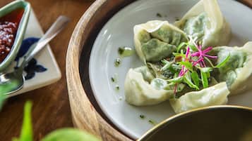 Um prato de dumplings decorado com ervas verdes e flores cor-de-rosa, acompanhado de uma pequena tigela de molho e uma colher.