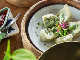 Um prato de dumplings decorado com ervas verdes e flores cor-de-rosa, acompanhado de uma pequena tigela de molho e uma colher.