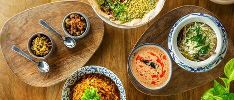 A variety of dishes on wooden serving platters, including a bowl of rice with green leaves, a bowl of curry garnished with cilantro, and a dish with pomegranate seeds and nuts.