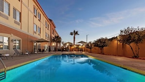 Ein Hotelpoolbereich mit Liegestühlen und Sonnenschirmen, umgeben von einem Gebäude mit vielen Fenstern und einer Palme.