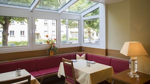 Een eetgedeelte met een zitbank, een tafel met tafelkleed, een lamp en uitzicht op de straat door grote ramen.