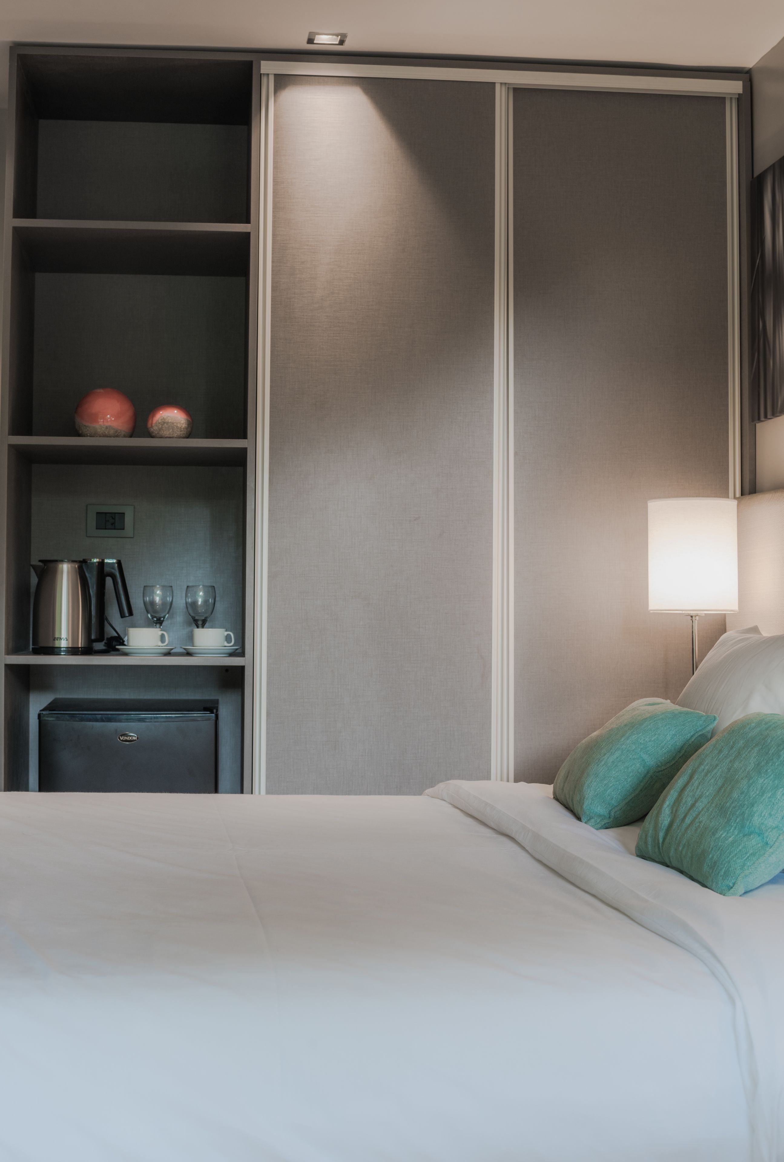 A hotel room with a bed, a minibar, a lamp, and a shelf with kitchenware.