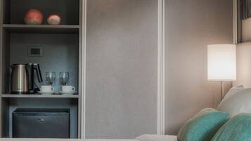 A hotel room with a bed, a minibar, a lamp, and a shelf with kitchenware.