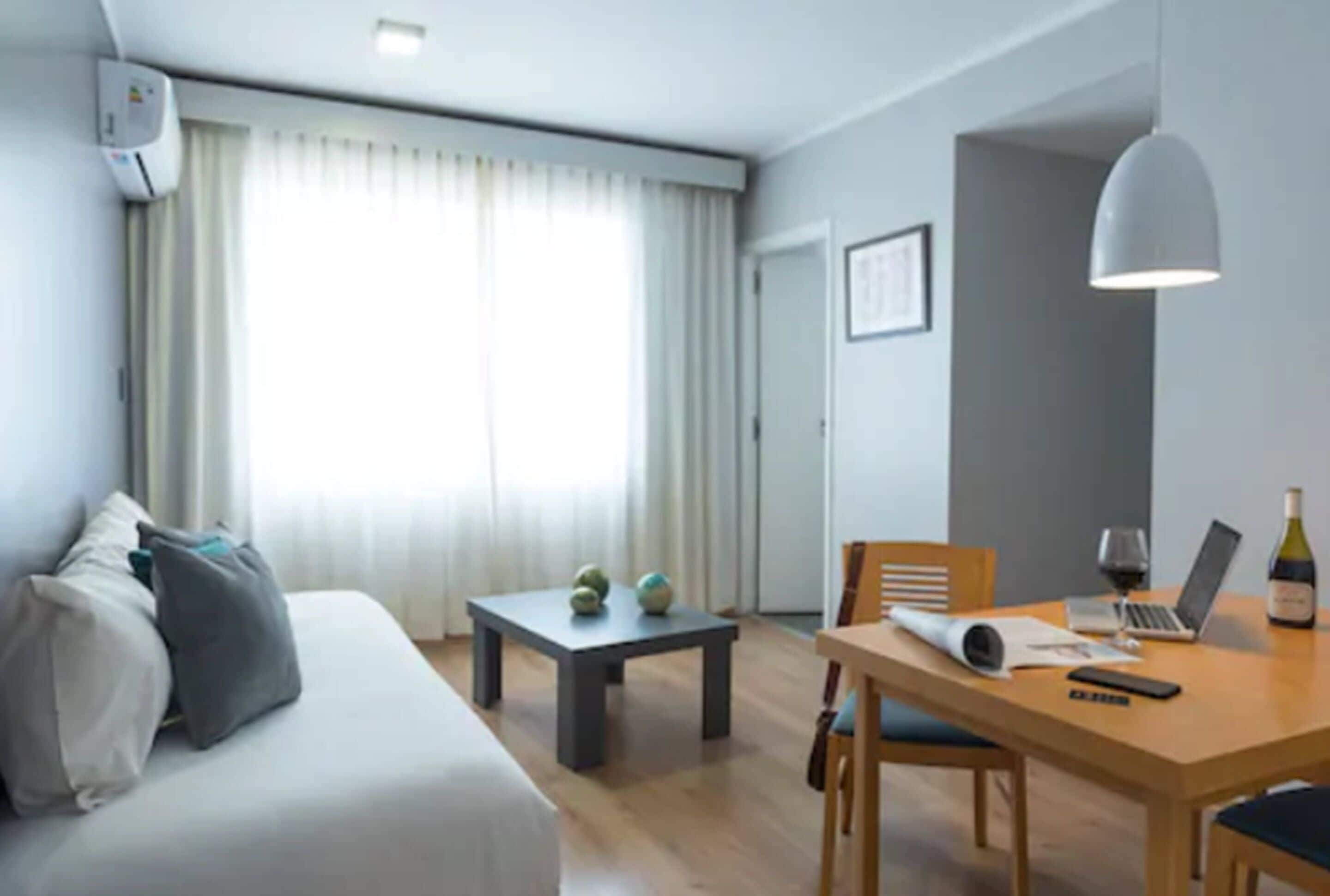 Et moderne hotelværelse med en hvid sofa, et lille bord med en bærbar computer og vin, samt et spisebord med stole.