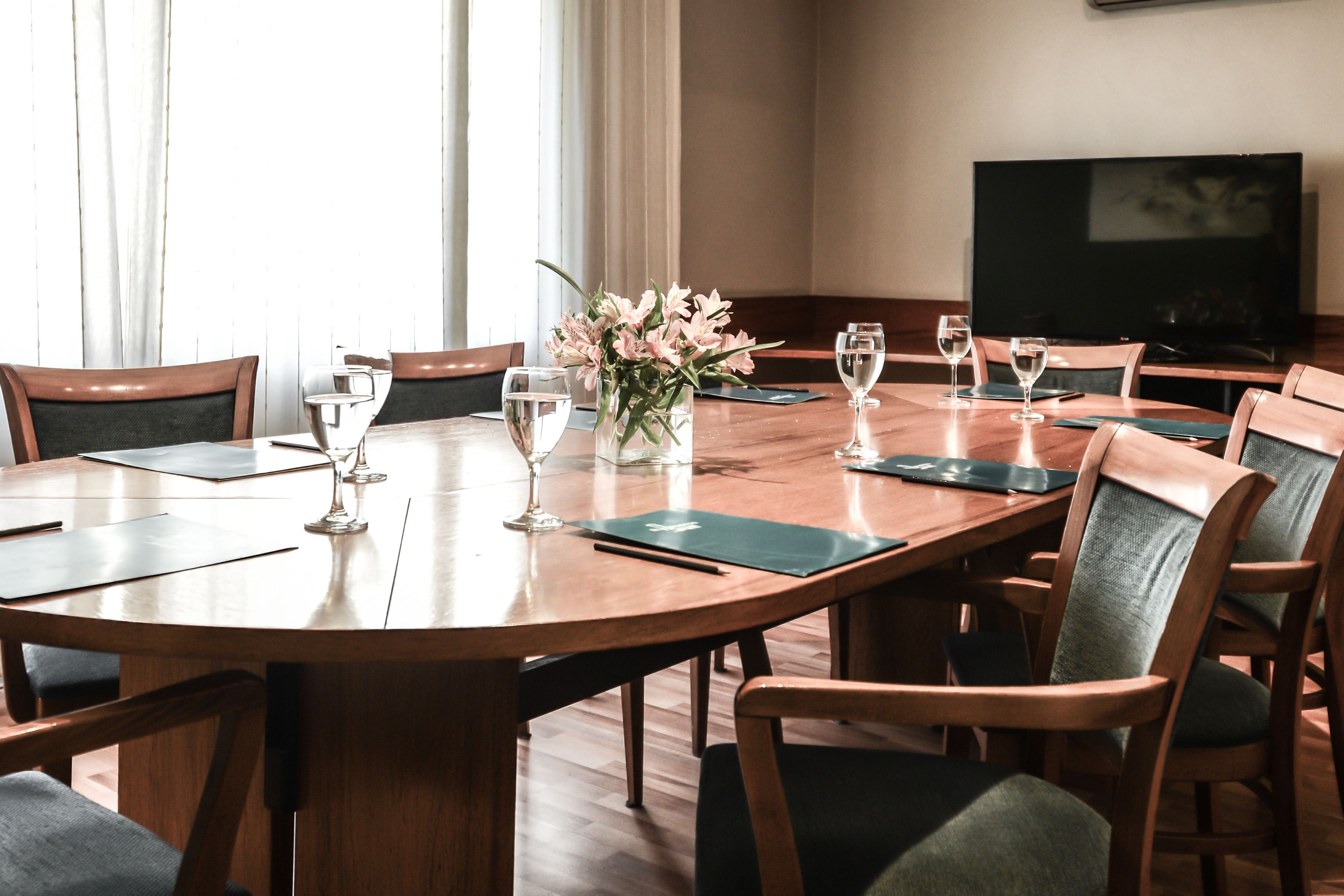 A conference room with a large oval table, wooden chairs, a vase of flowers, and a television on the wall.
