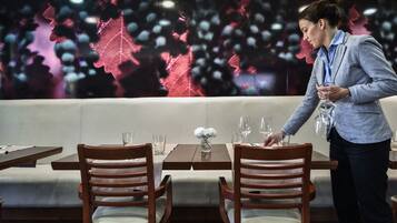 Una zona de comedor con una mesa dispuesta para dos, un florero con flores y una persona ordenando vasos.