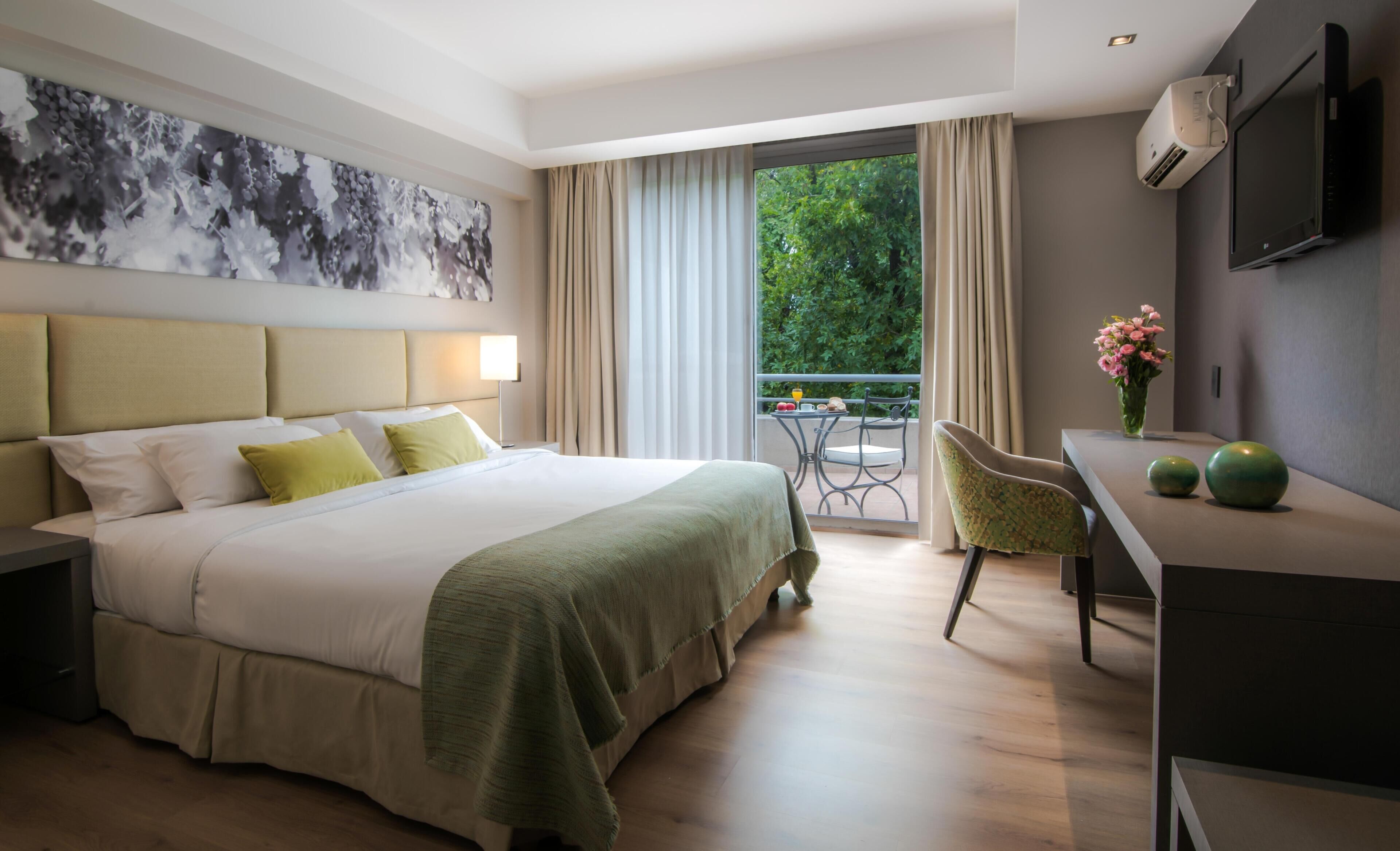 A hotel room with a bed, a desk, a chair, a TV, and a balcony with a view of greenery.